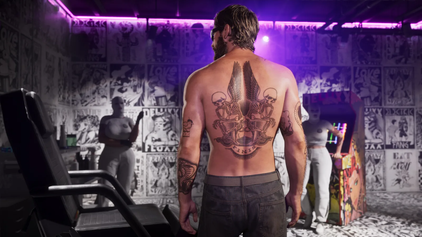 A shirtless man seen from behind showing a large winged-skeleton back tattoo lettered FINIS, standing in a studio papered floor to ceiling with black-and-white flash art, two masked staff and an arcade cabinet behind him.
