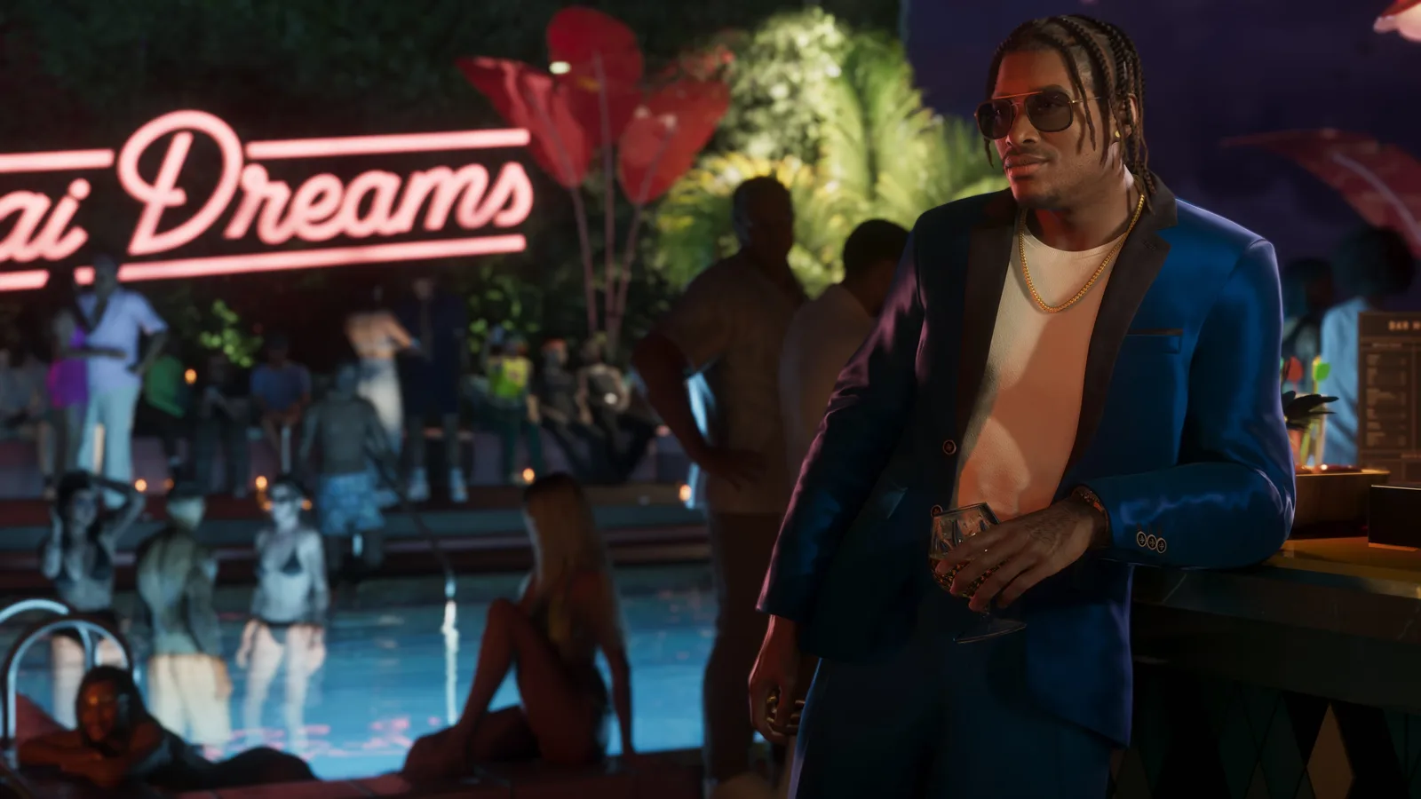 DreQuan Priest in a blue satin blazer and gold chain holding a drink at an outdoor bar beside a floodlit pool, a neon sign glowing over the crowd behind him.
