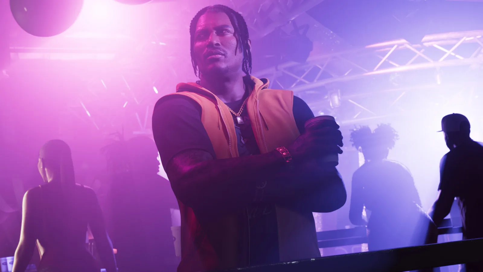 DreQuan Priest leaning on a rail with a drink in a hooded orange gilet under pink and violet stage lighting, silhouetted club-goers and a lighting truss behind him.