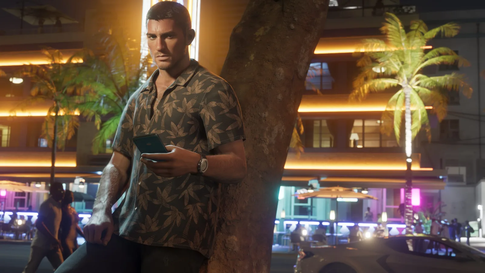 Jason Duval leaning against a tree at night in a dark floral shirt, holding a phone, on a street lit by neon and the amber lights of a building behind him.