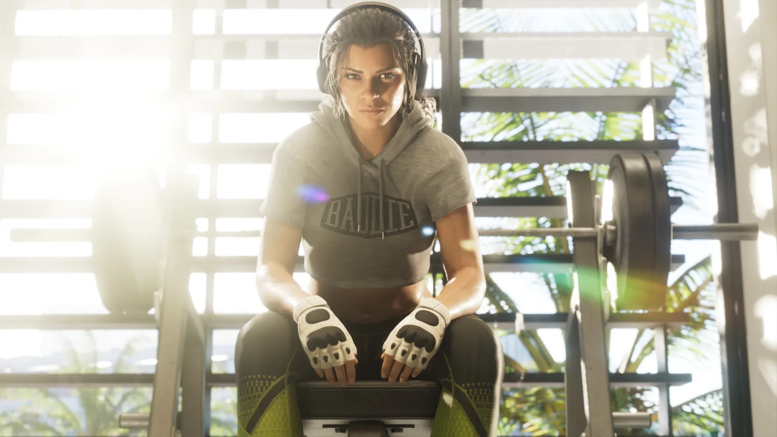 Lucia Caminos sitting on a weight bench in a sunlit gym in a cropped hoodie, headphones and fingerless training gloves, a loaded barbell rack beside her.