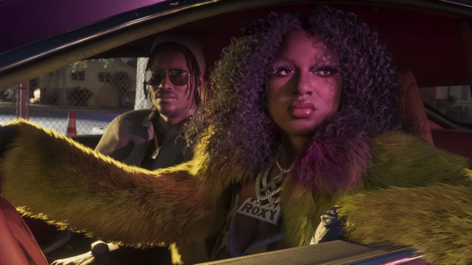A member of Real Dimez with purple curls in a yellow fur coat and a large pendant sitting in a car's passenger seat, a man in sunglasses and a cap at the wheel beside her.