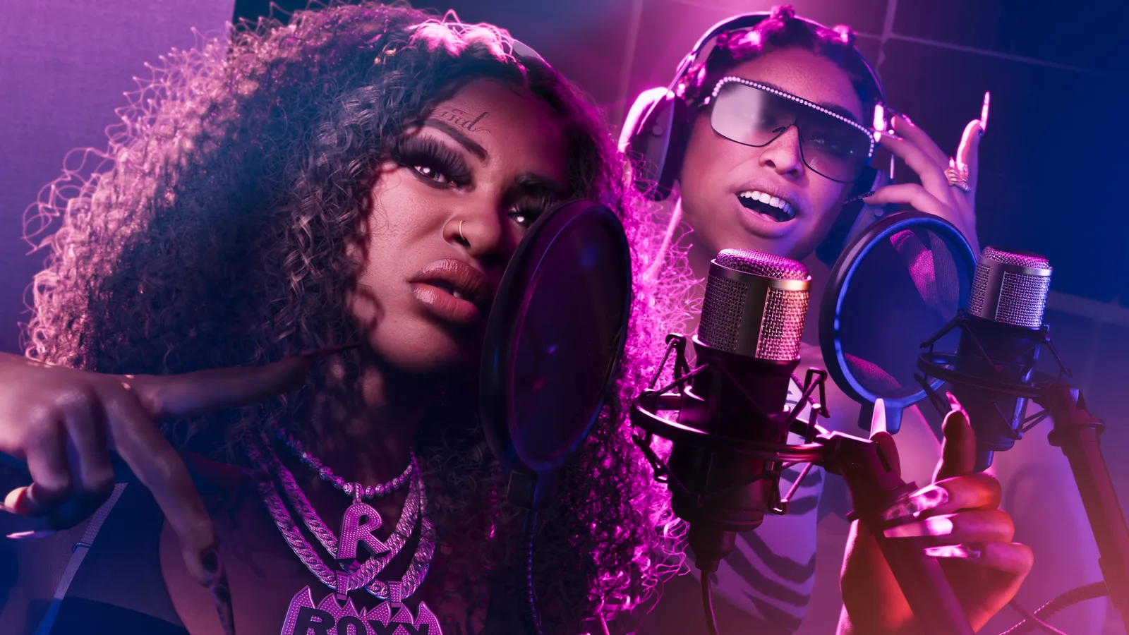 The two members of Real Dimez recording at studio microphones behind pop filters under magenta light, one in a diamond chain and one in headphones and rhinestone shades.