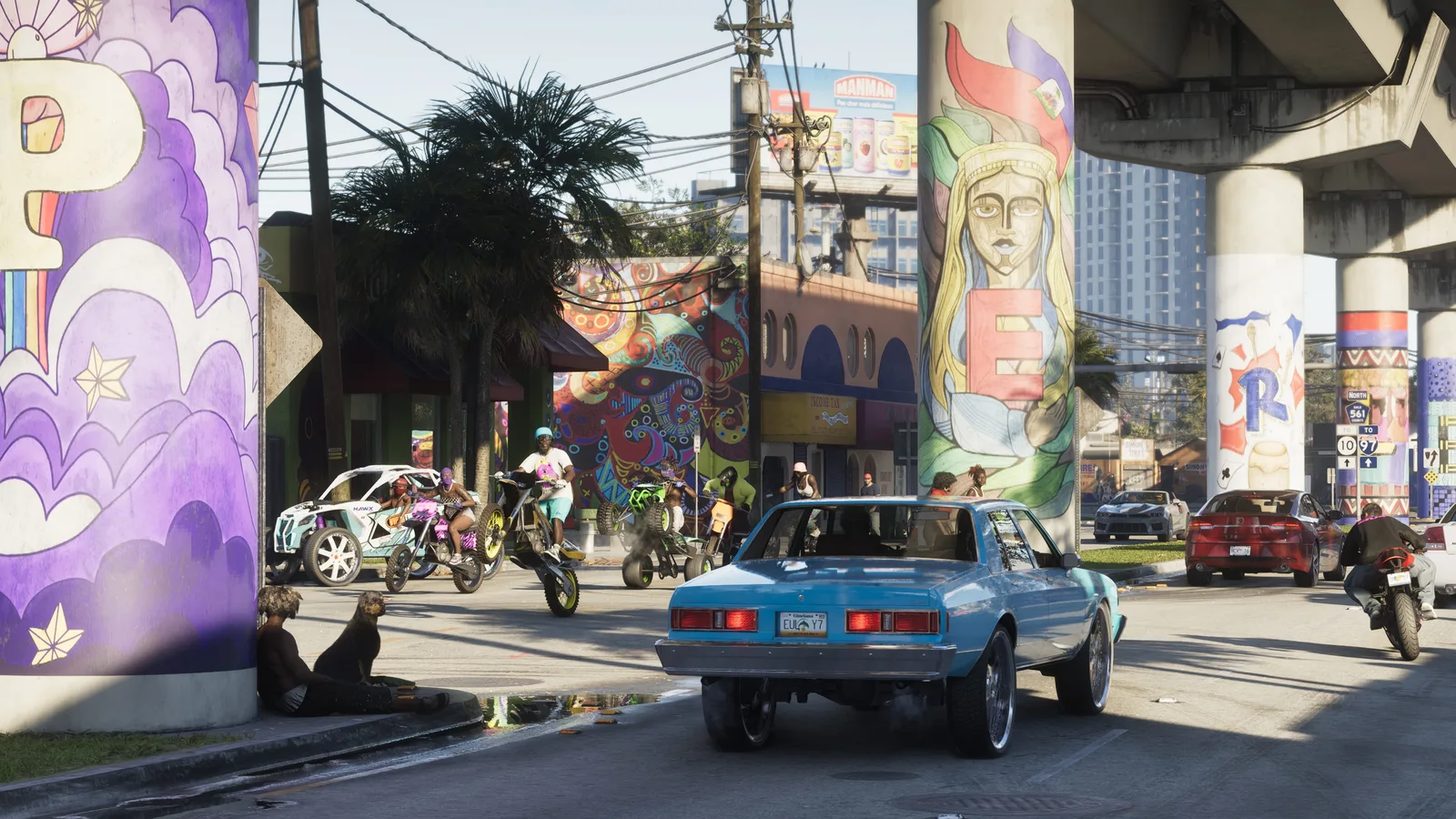 A daylight street beneath an elevated highway whose columns are painted with murals, a blue classic sedan on chrome wheels in the foreground and riders on dirt bikes, a quad and a buggy gathered at the kerb.