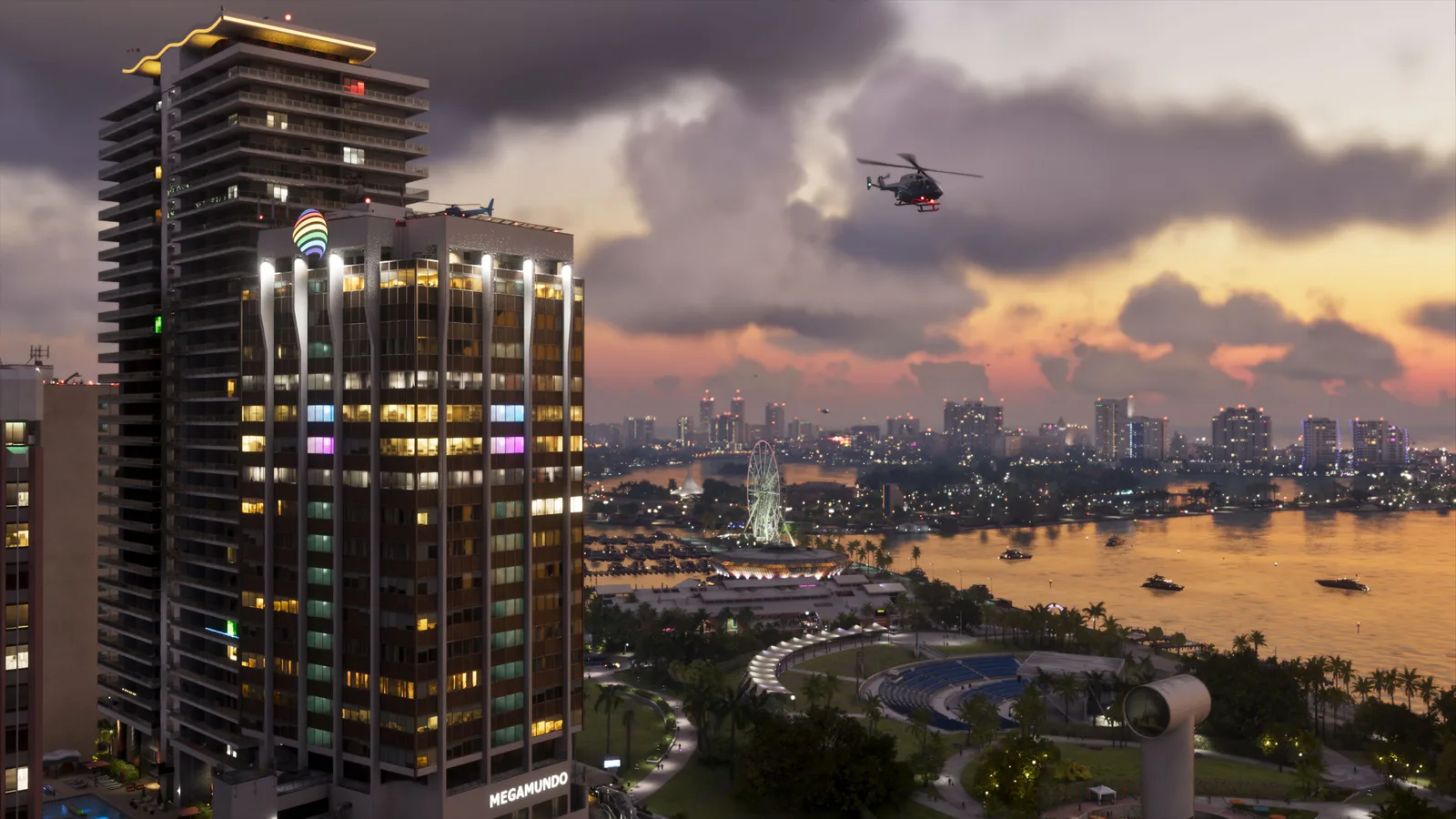 A high view at dusk of a lit Megamundo tower beside a bayfront park and amphitheatre, a Ferris wheel and marina below, a helicopter overhead and the far shore glowing across the water.