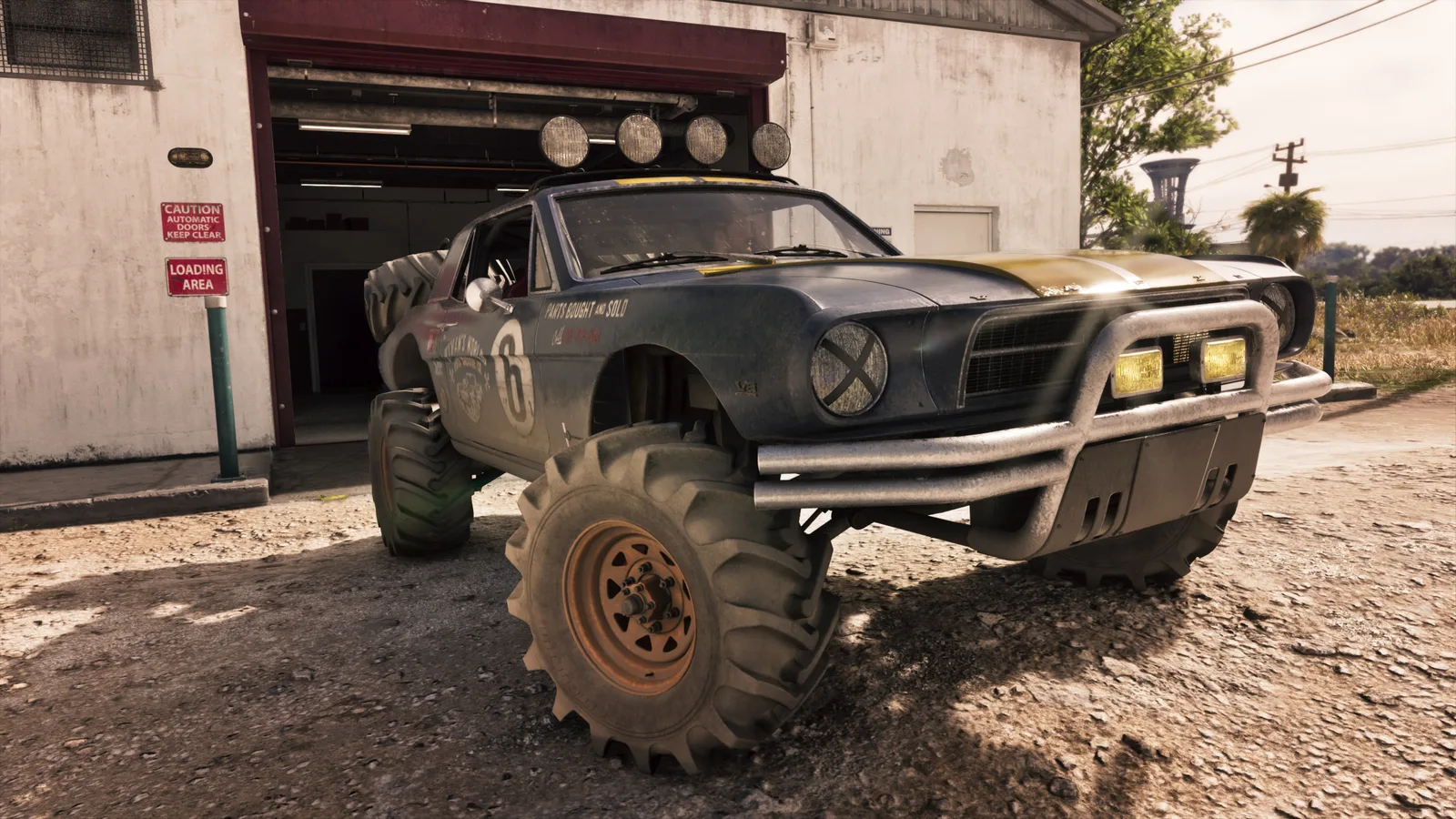 The blue and yellow buggy parked on gravel outside a workshop loading bay on huge tractor-tread tyres, a roof light bar above and a race number 6 and PARTS BOUGHT AND SOLD lettering on its flank.